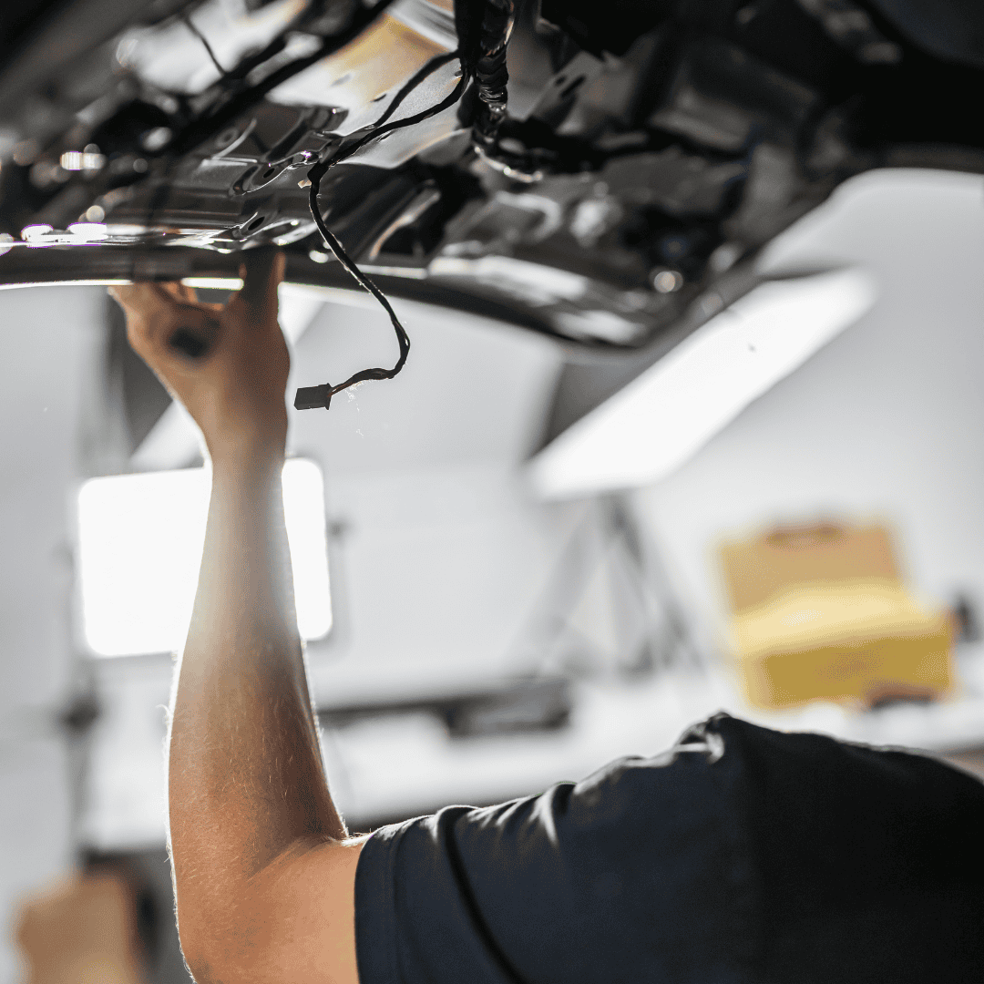 Dirbtuvėse, po ryškiomis šviesomis, žmogus dirba su automobilio bagažine, reguliuodamas laidus. Ši aplinka perteikia susitelkimą ir techninius įgūdžius.