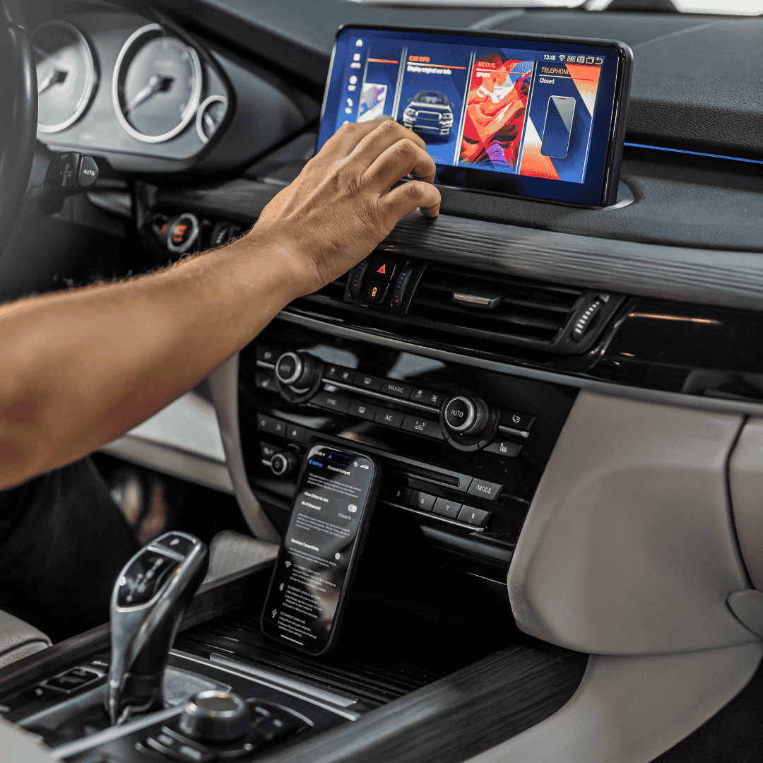 Ranka sąveikauja su automobilio liečiamu multimedijos ekranu, kuriame virš prietaisų skydelio valdiklių rodomos programėlės. Išmanusis telefonas padėtas prie multimedijos. Scena perteikia šiuolaikines technologijas.