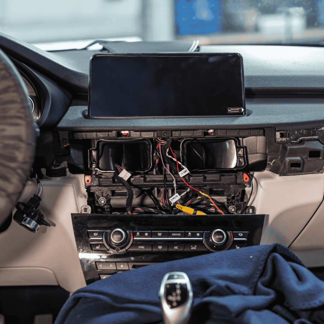 Automobilio prietaisų skydelis su nuimtu skydeliu, kuriame matyti laidai ir komponentai. Viršuje sumontuotas didelis multimedijos ekranas.